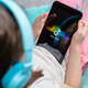 A young girl with blue headphones lays back on a chair cradling a tablet with the TikTok logo on it.